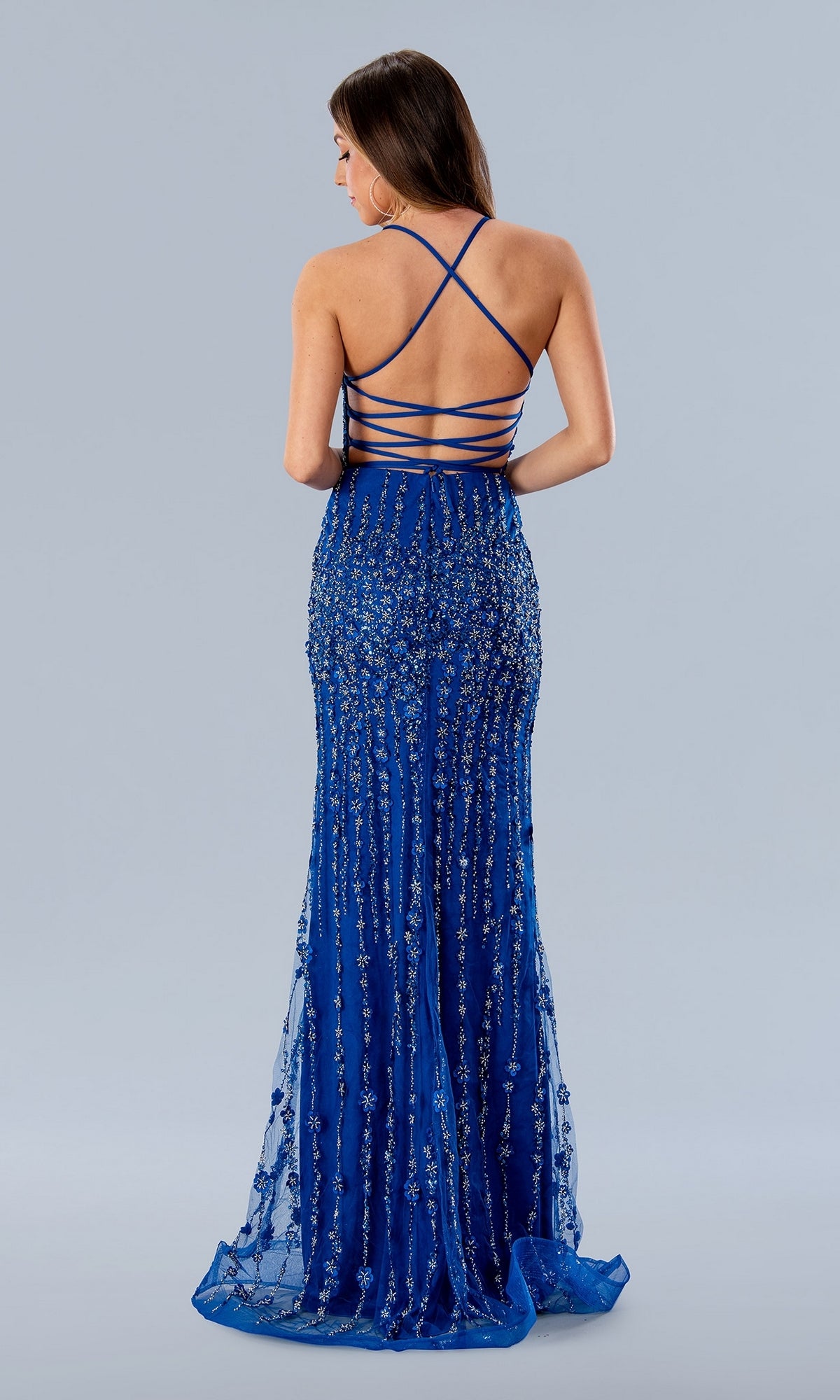 A woman stands facing away, wearing the Long Prom Dress 24189 by Stella Couture—a fitted royal blue gown with sequin embellishments and crisscross spaghetti straps over an open lace-up back.